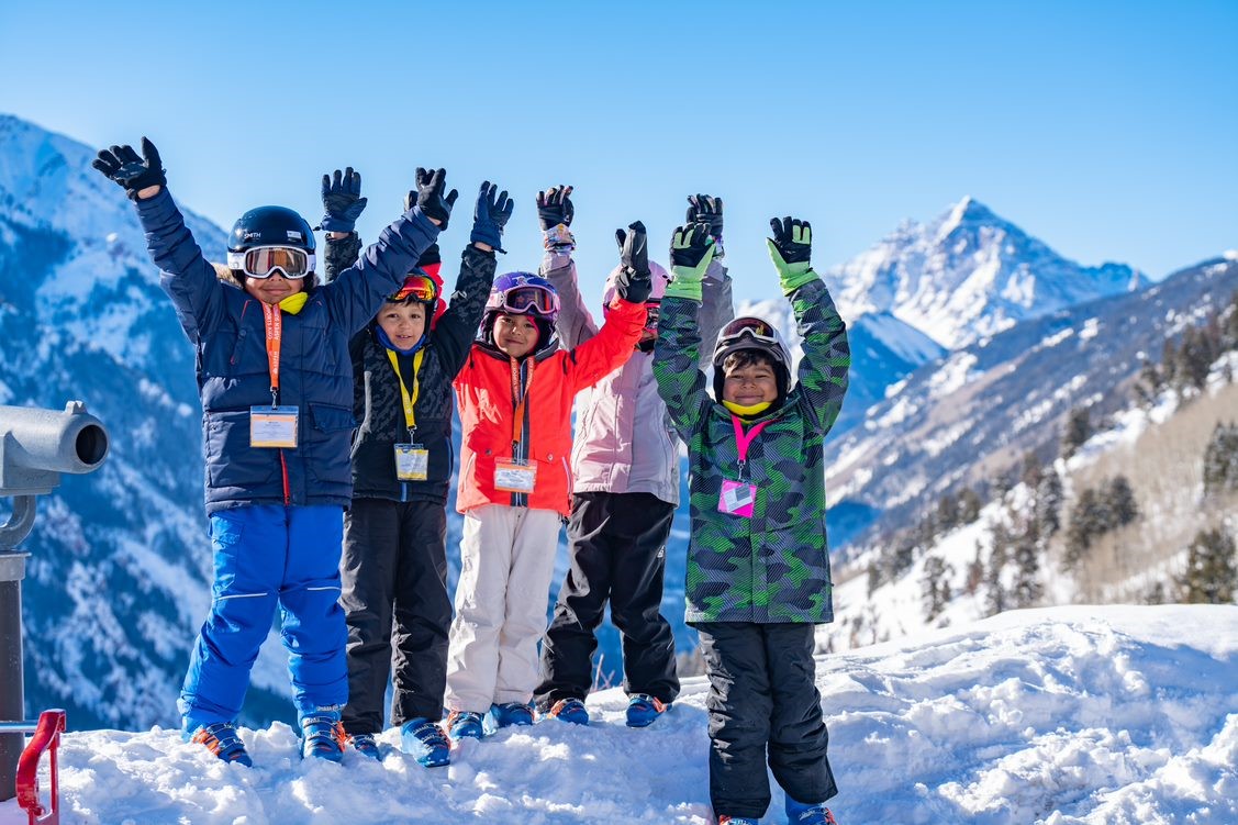 A group of 5 young children in ski gear standing on a snowy hill.