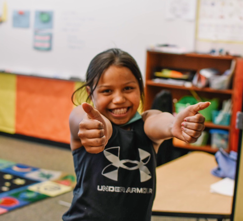 A young girl of color smiling and giving two thumbs up in a classroom.