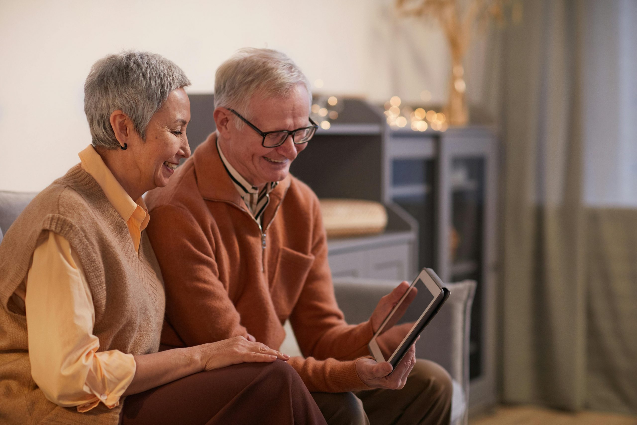 Two white elderly individuals sitting side by side on a couch and smiling at a tablet device.